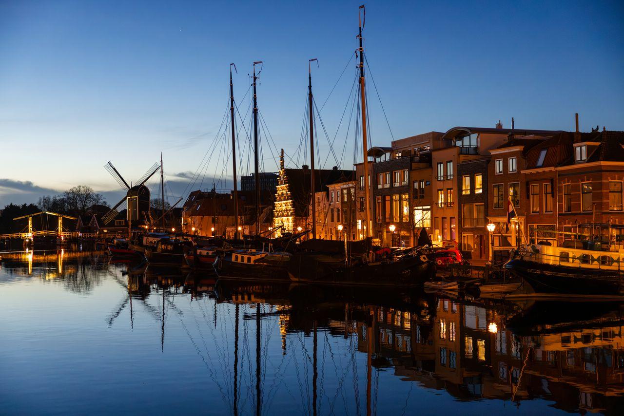 Historic sailing barges moored along a Leiden canal at dusk, with a windmill and lit houses on the quay
